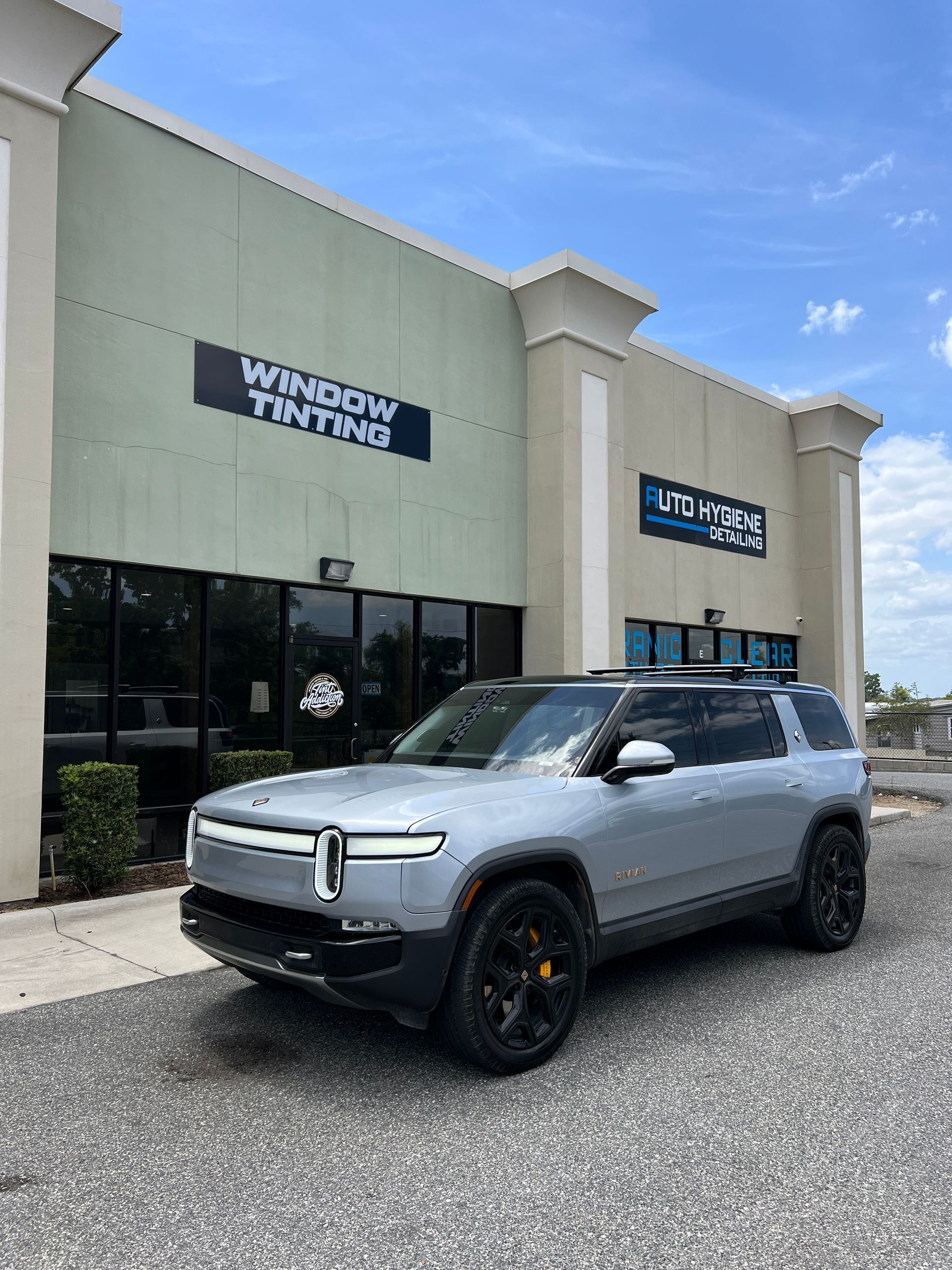 Silver Rivian SUV parked in front of a window tinting business with a blue sky.
