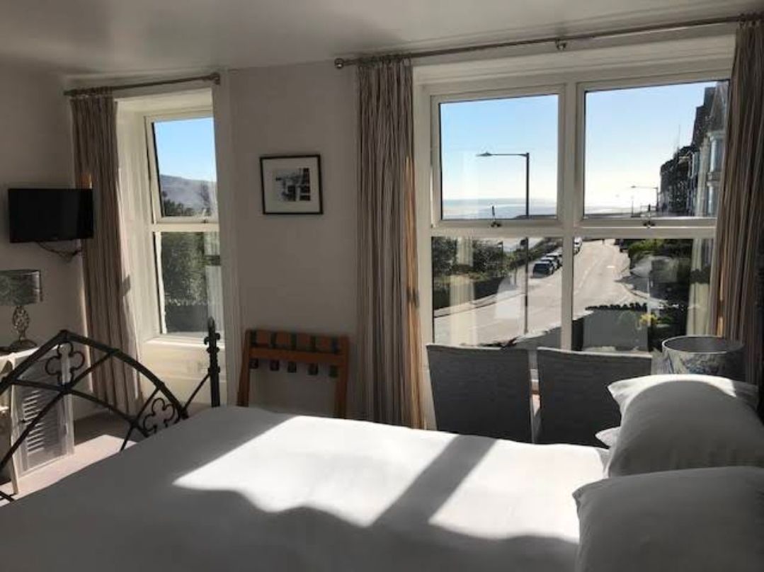 Bright bedroom with large windows, a bed, and sunlight streaming in toward a balcony and street view.