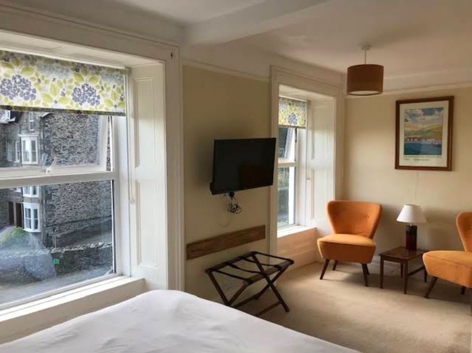 Bright hotel room with two windows, wall-mounted TV, orange chair, and bed in foreground