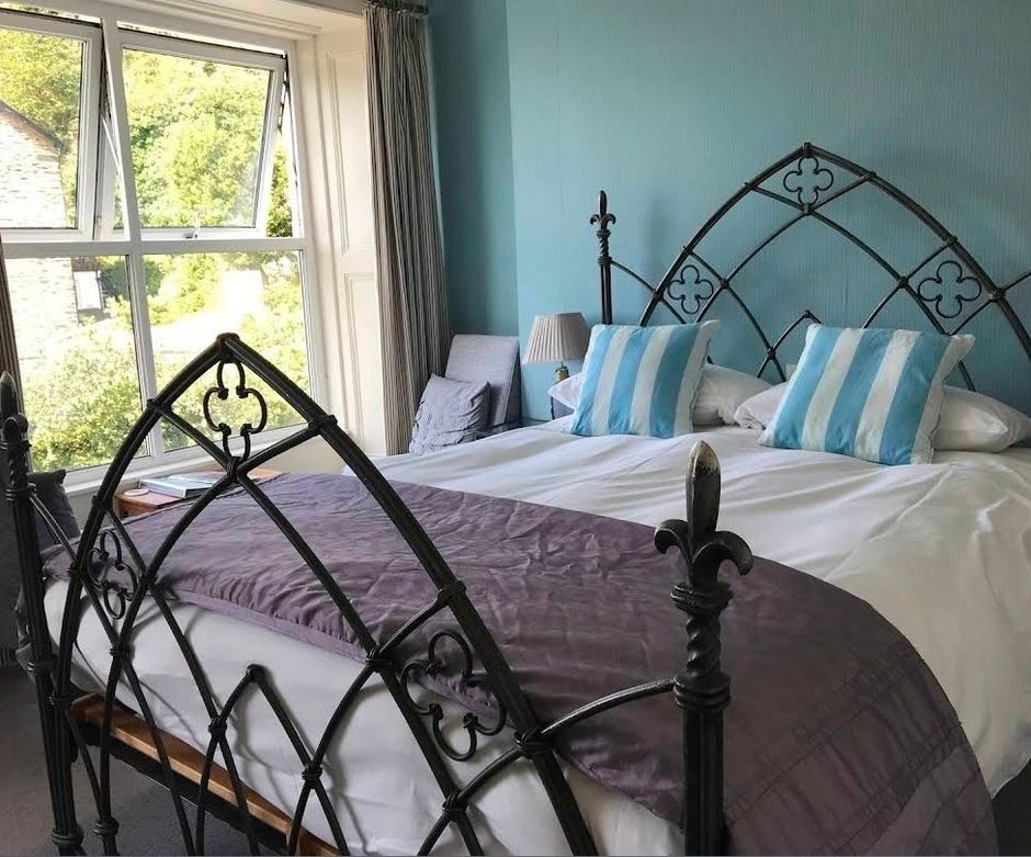 Bright bedroom with wrought-iron bed, white bedding, blue pillows, and a window overlooking greenery.