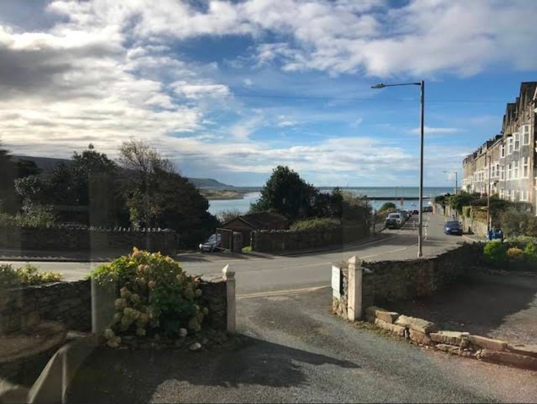 Seaside street with stone walls, parked cars, hillside buildings, and a view of the bay under cloudy skies