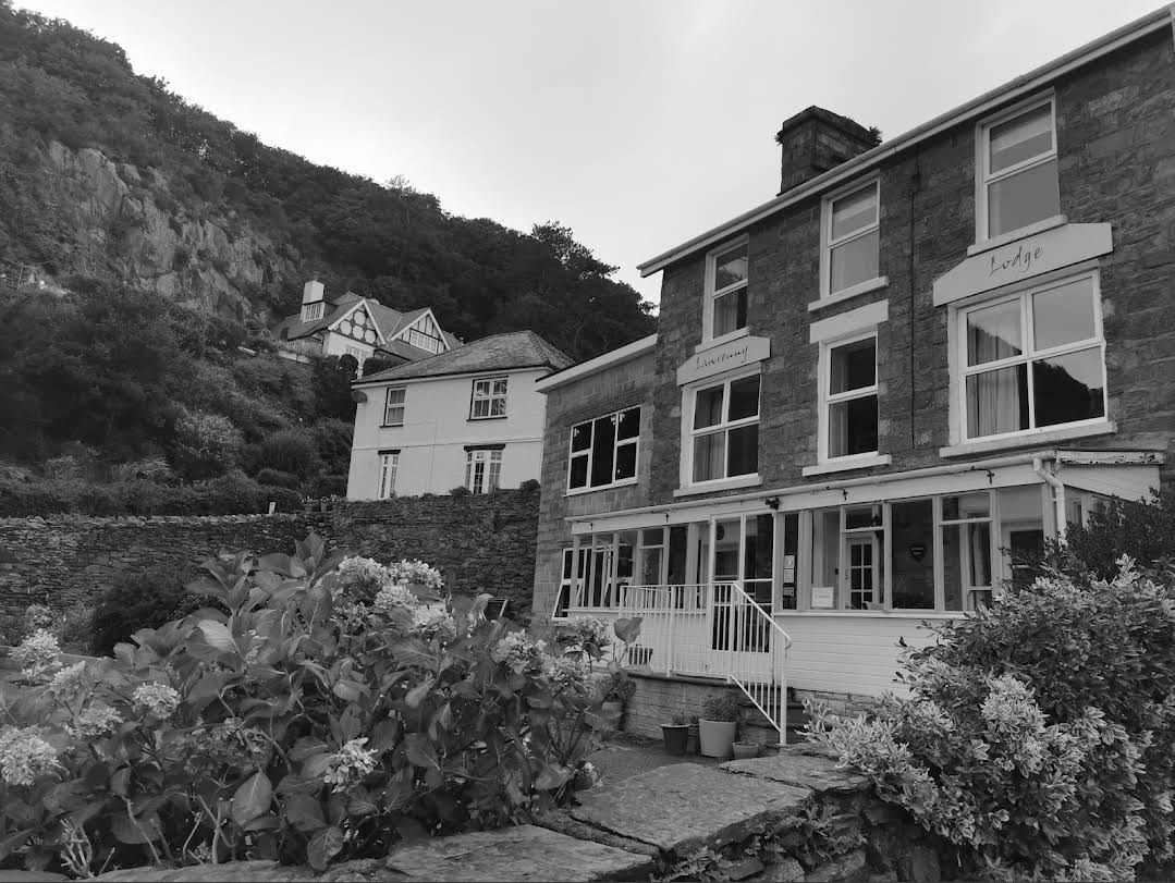 Stone cottage with white trim beside a garden, set against a steep wooded hillside.