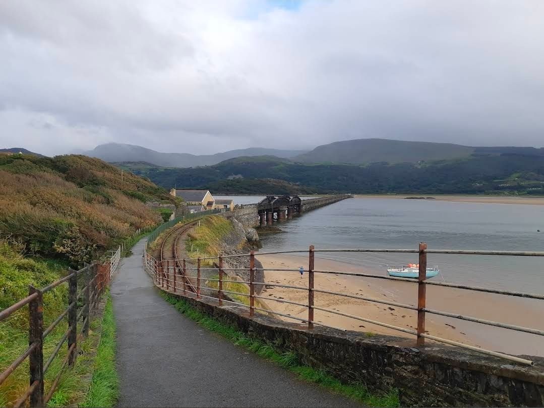 Coastal path beside a beach and bay under cloudy skies, with hills in the distance
