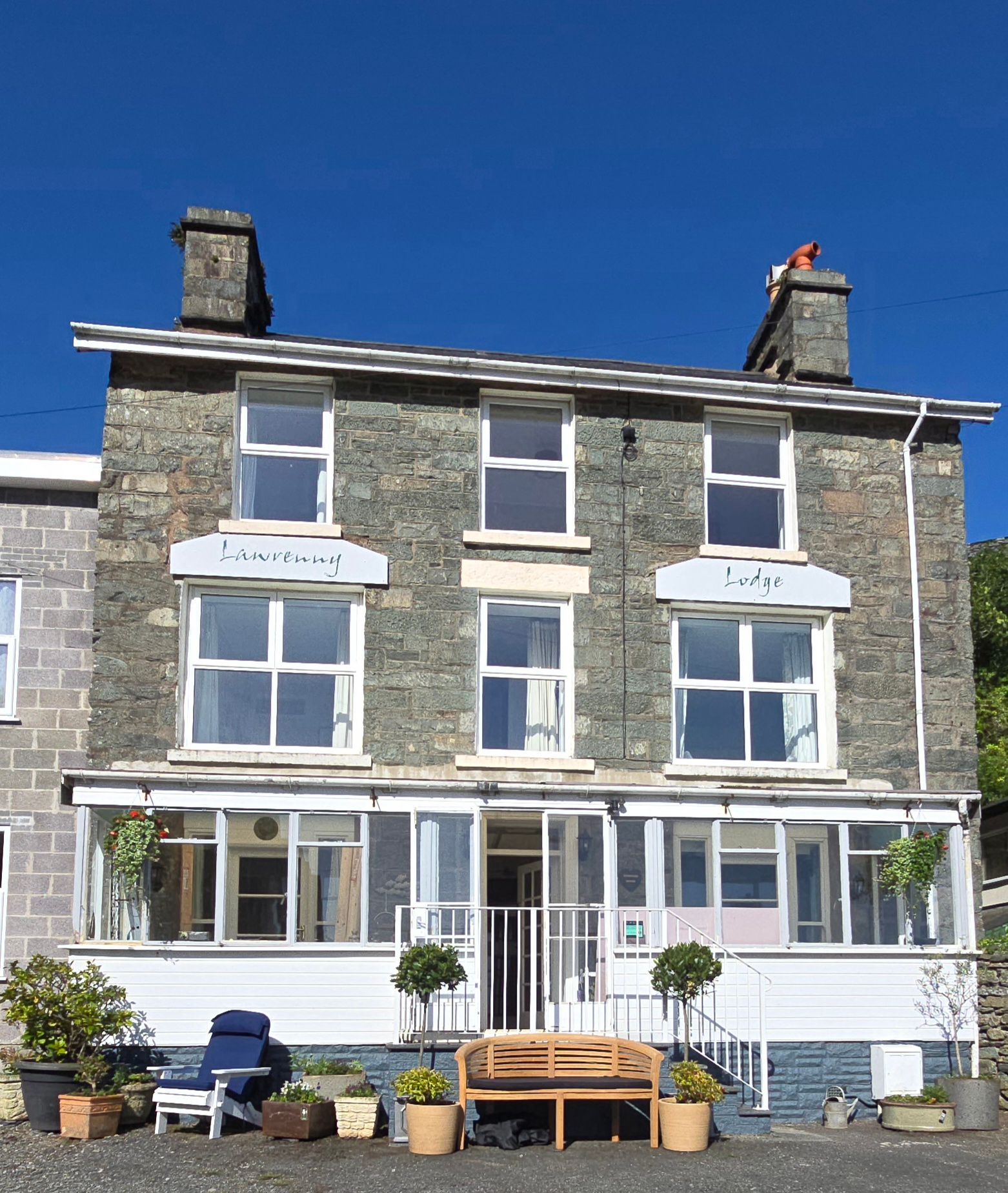 Stone seaside house with white-trimmed windows, balconies, and potted plants under a clear blue sky