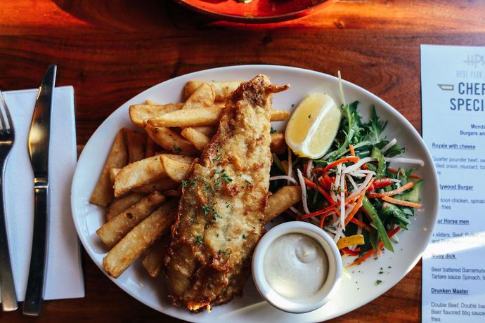 Plate of fried fish, thick-cut chips, coleslaw, lemon wedge, and tartar sauce on a wooden table.