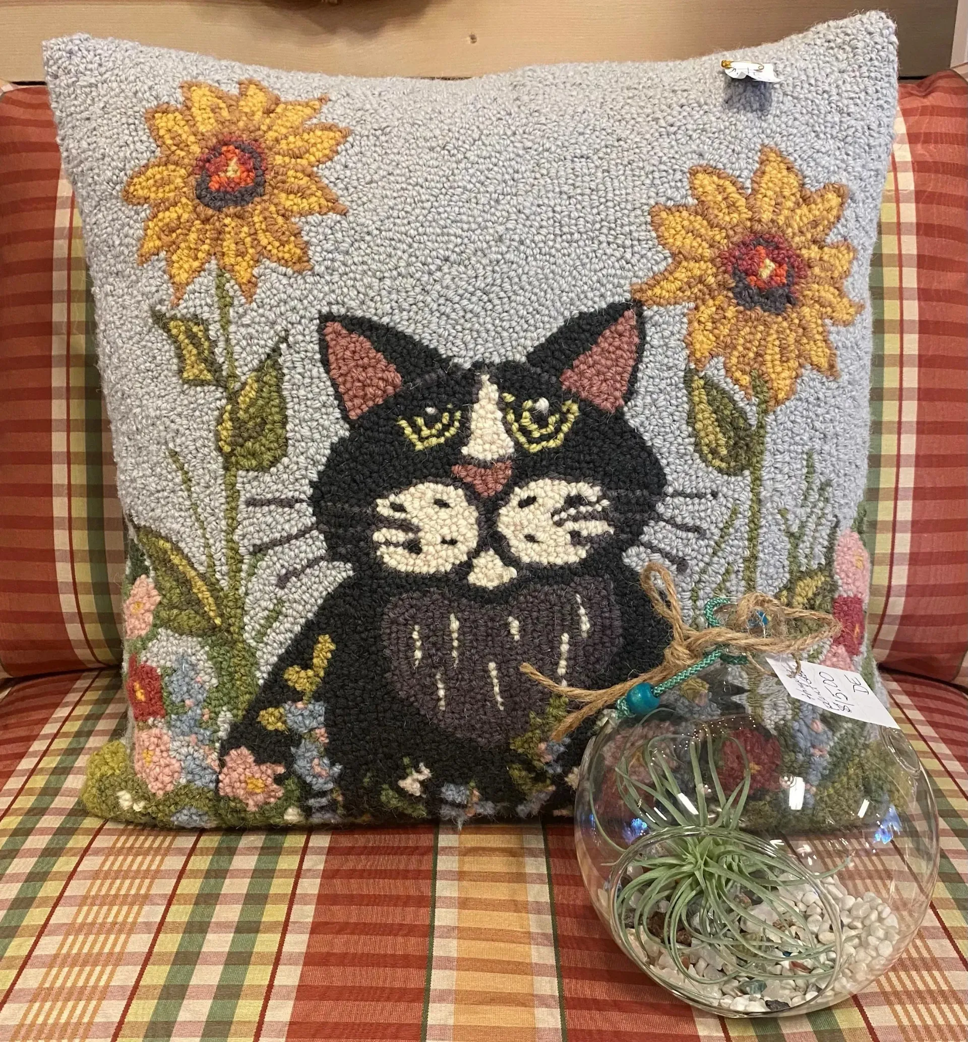 Cat tapestry pillow with yellow sunflowers beside a small glass terrarium on a plaid couch