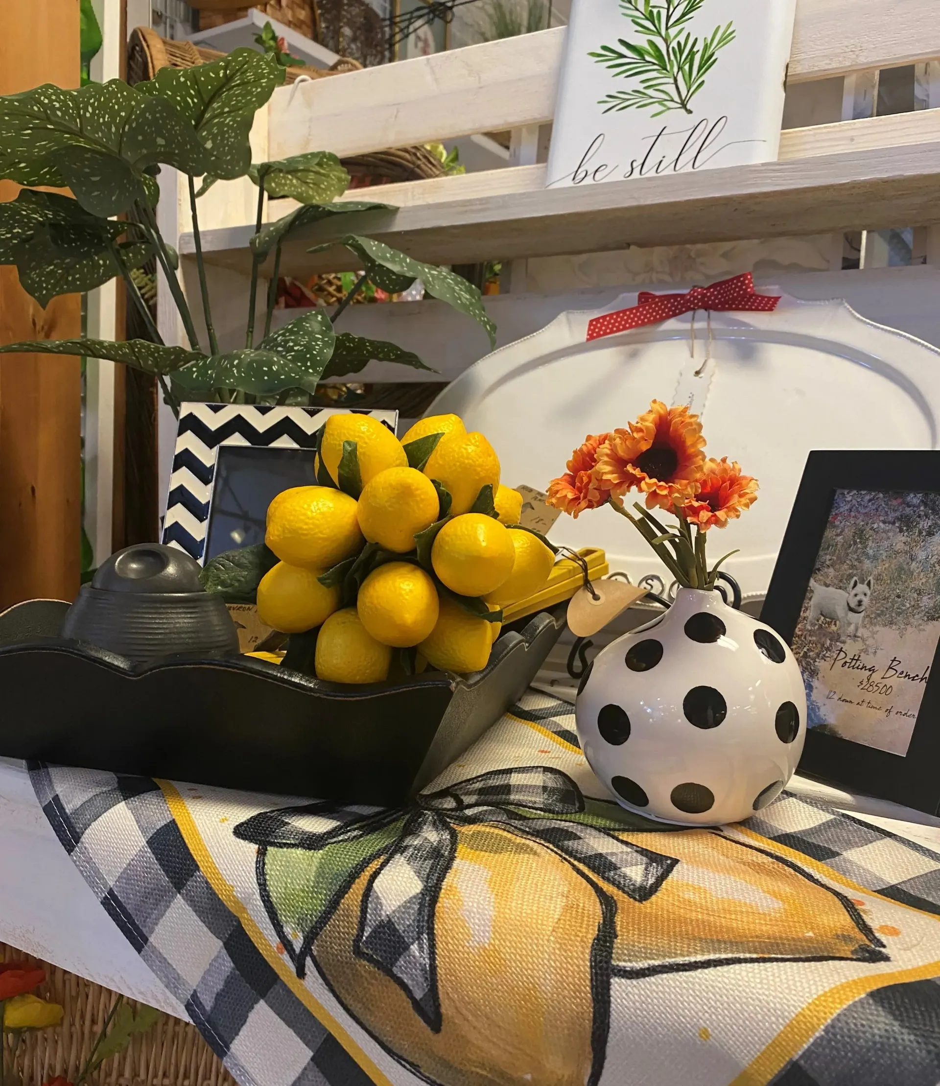 Decorative shelf with lemons, flowers in a polka-dot vase, and framed art on a patterned cloth