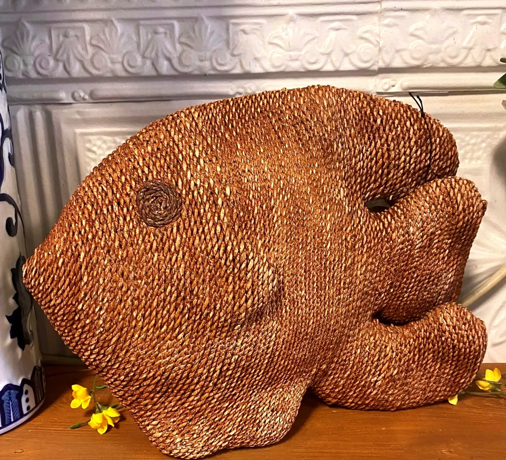 Brown woven fish-shaped sculpture on a wooden surface, with small yellow flowers nearby.