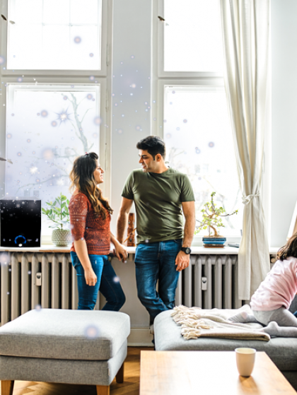 Two people talking by a bright window in a cozy living room with a couch and radiator nearby