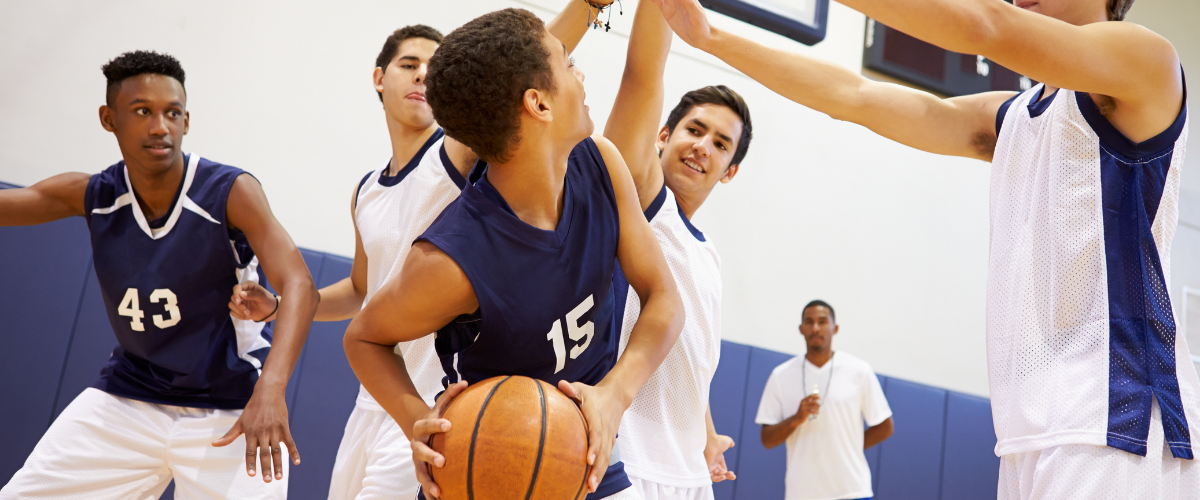 Basketball players in a gym, one with the ball being blocked by an opponent, other players look on.
