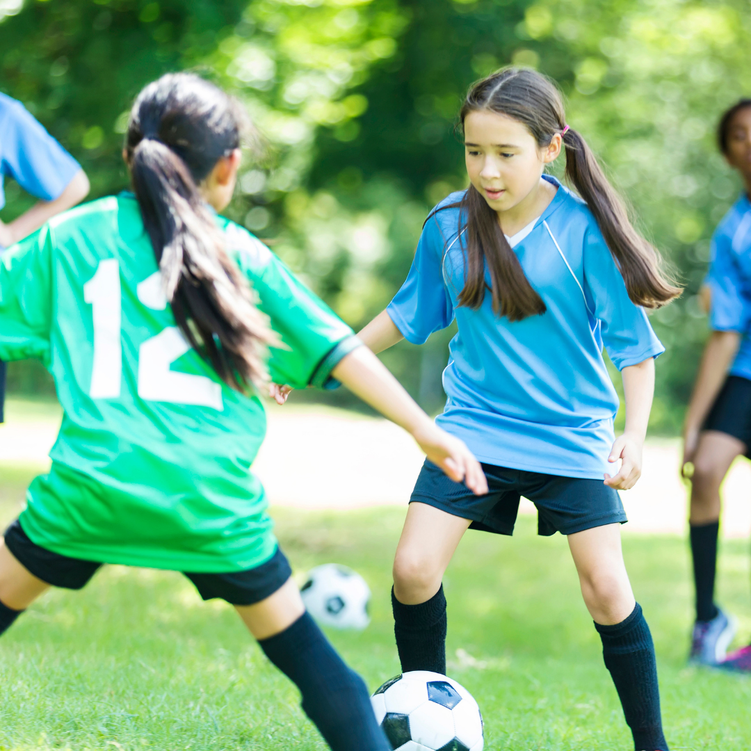 Two young girls playing soccer on a green field; one in green jersey, the other in blue, with soccer ball.