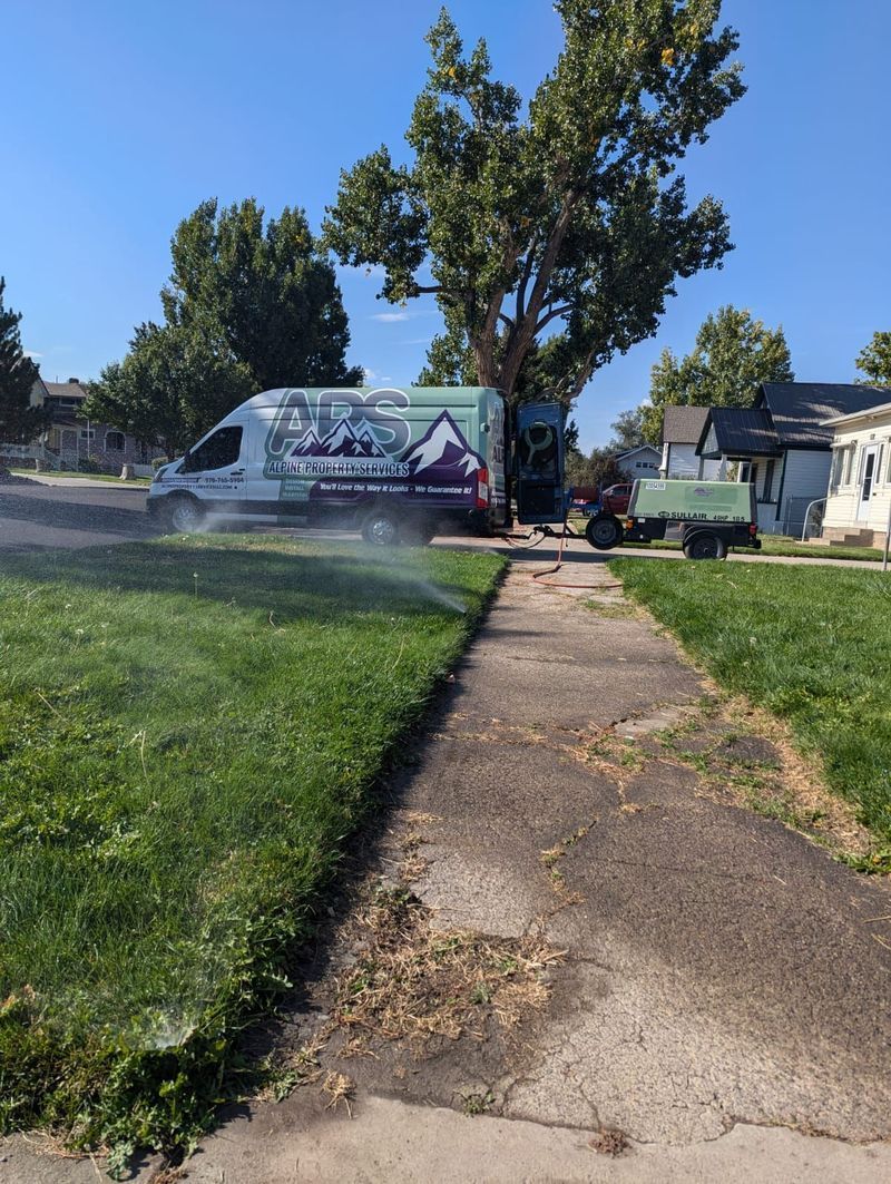 A van parked on a street has a mountain and forest graphic. A sprinkler waters the grass beside a sidewalk.