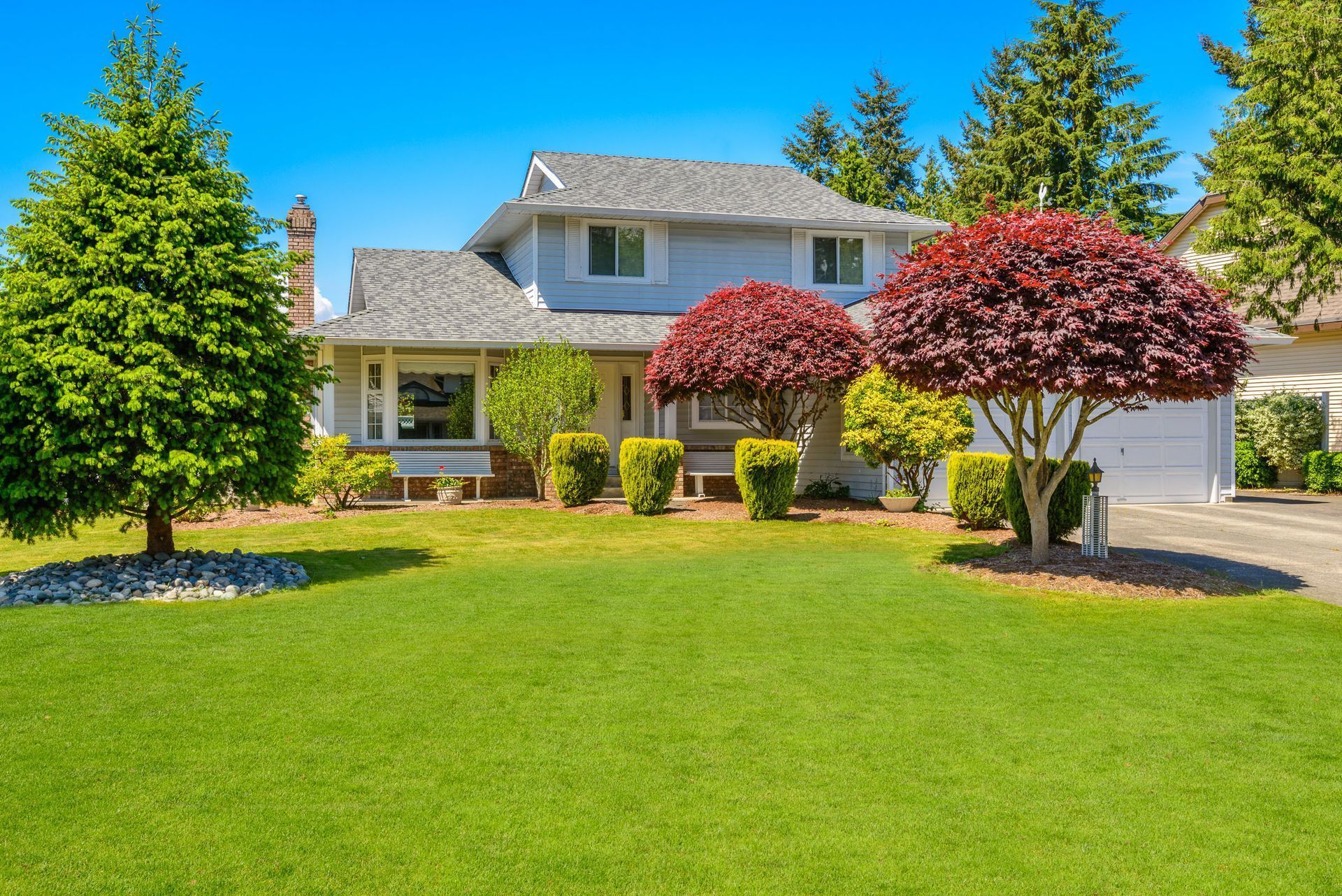 Two-story blue house with green lawn, shrubs, trees, and driveway on a sunny day.