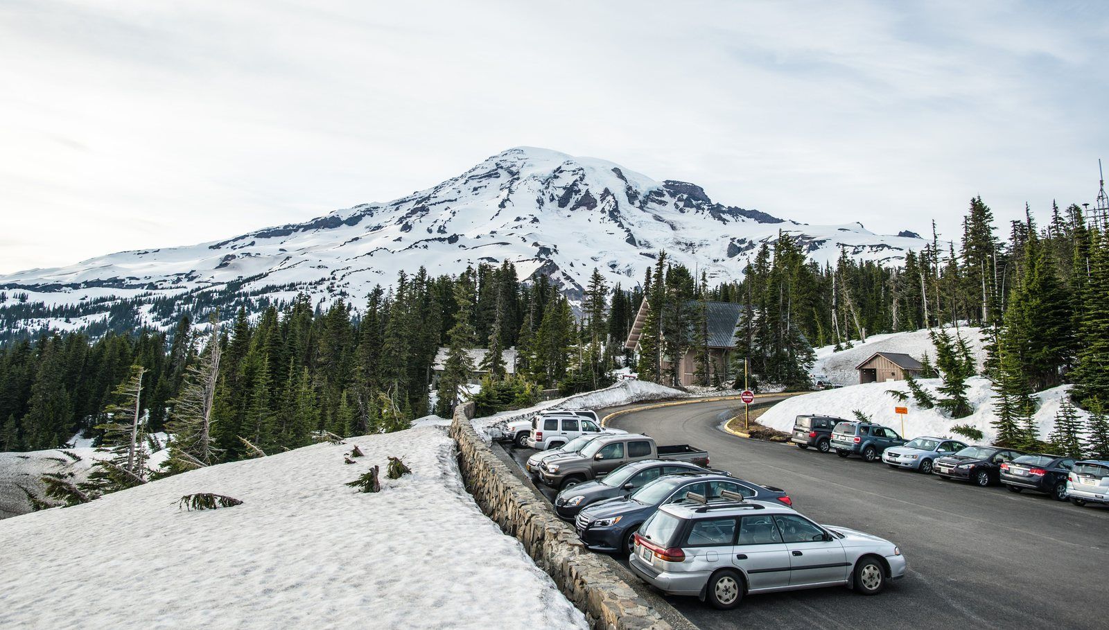 Cleared mountain parking lot with cars parked beside plowed snow banks below a snow-capped peak