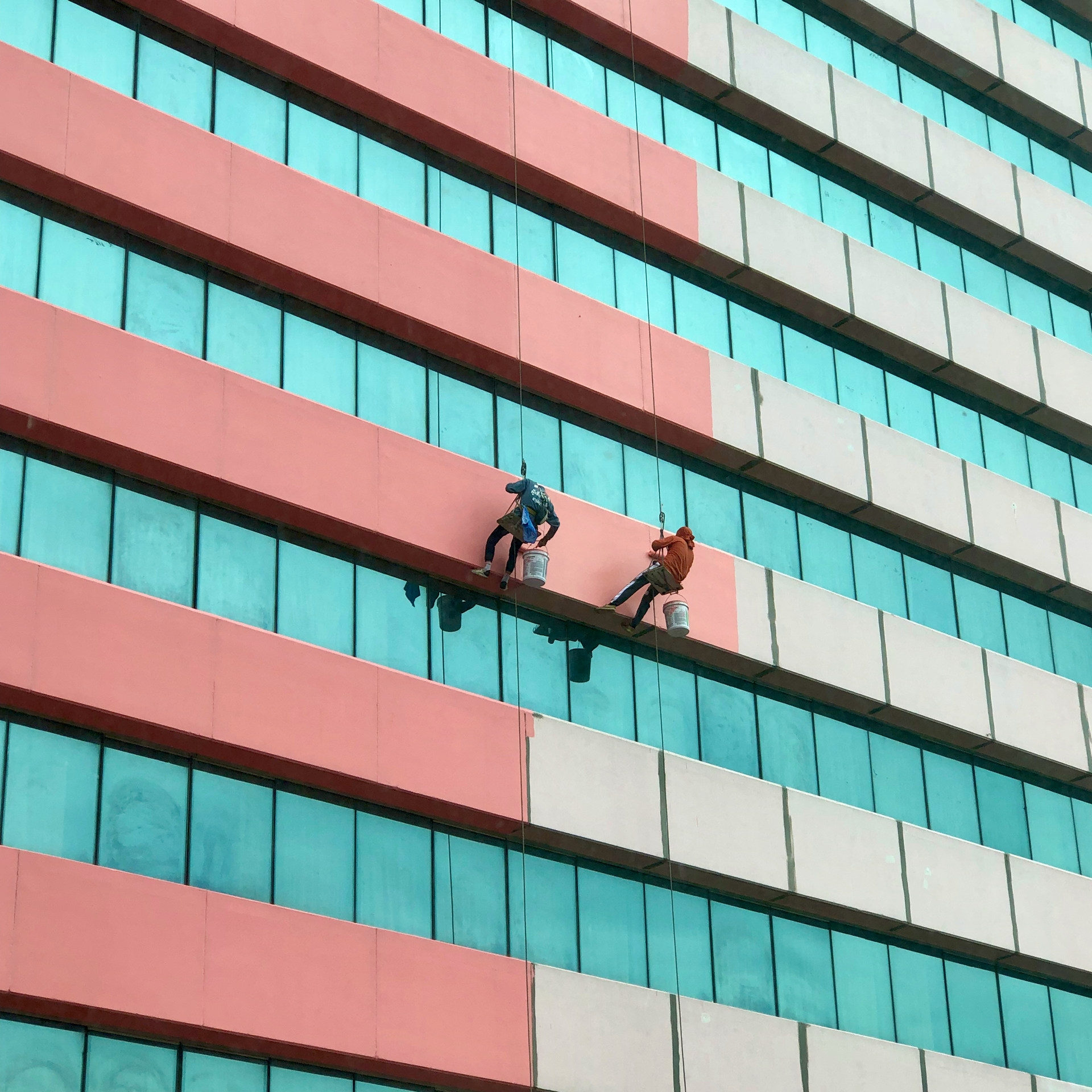 Two workers on ropes cleaning a turquoise and coral high-rise facade
