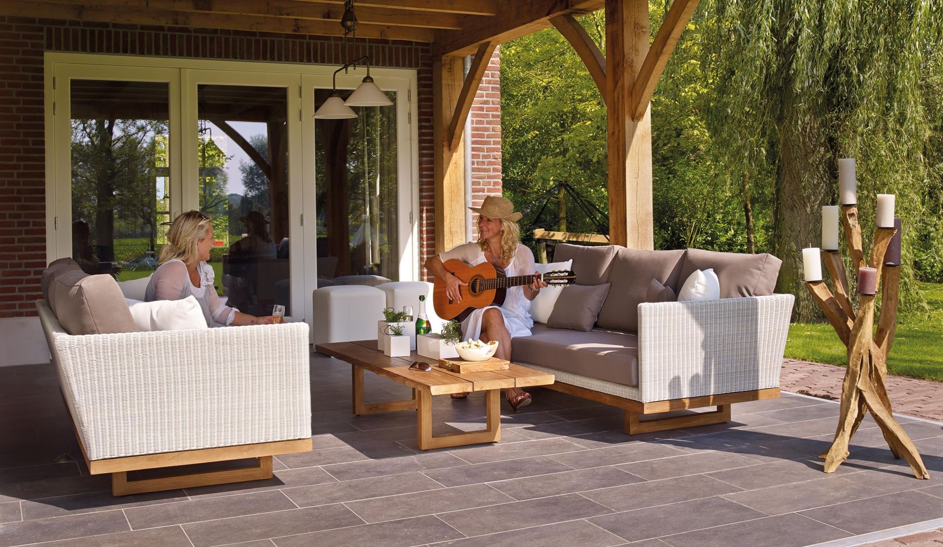 Outdoor patio with wicker sofas, coffee table, and a person playing guitar under a wooden pergola.