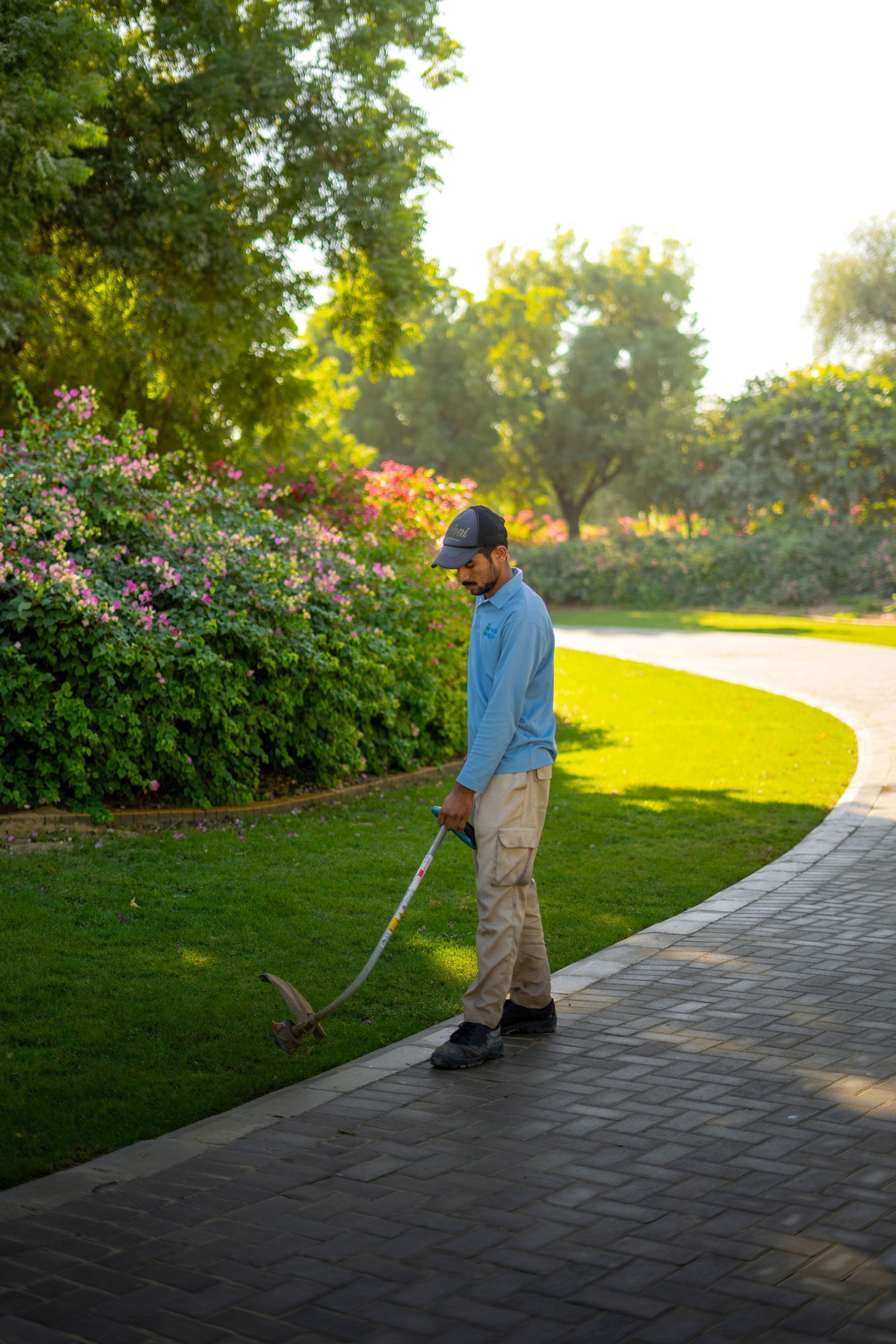 Man using a golf club to practice on a curved path beside a sunny garden with flowers and trees