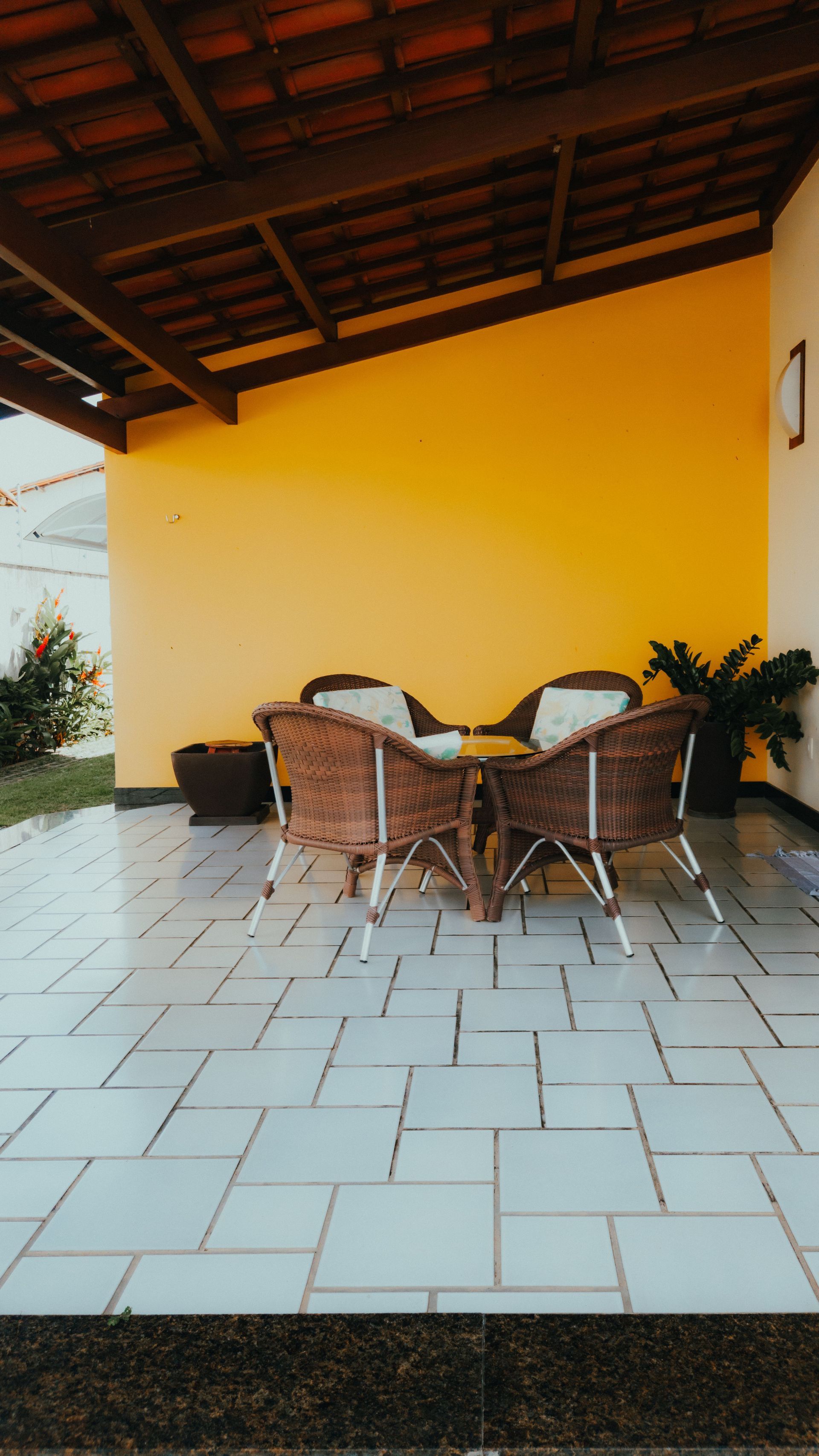 Covered patio with wicker dining set on a tiled floor, against a yellow wall and wooden roof.