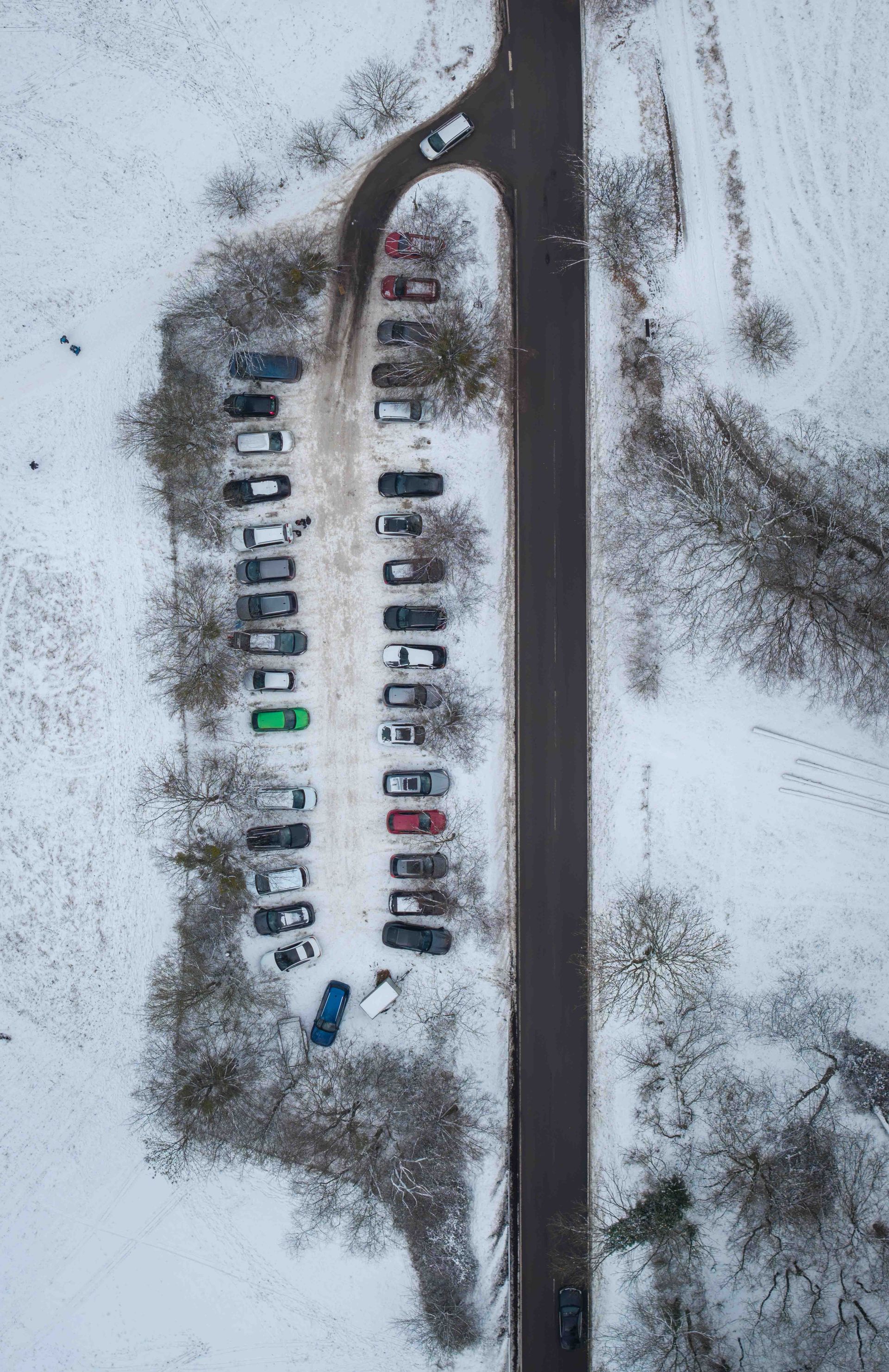 Aerial view of a snowy parking lot with rows of parked cars beside a long road and a curving driveway.