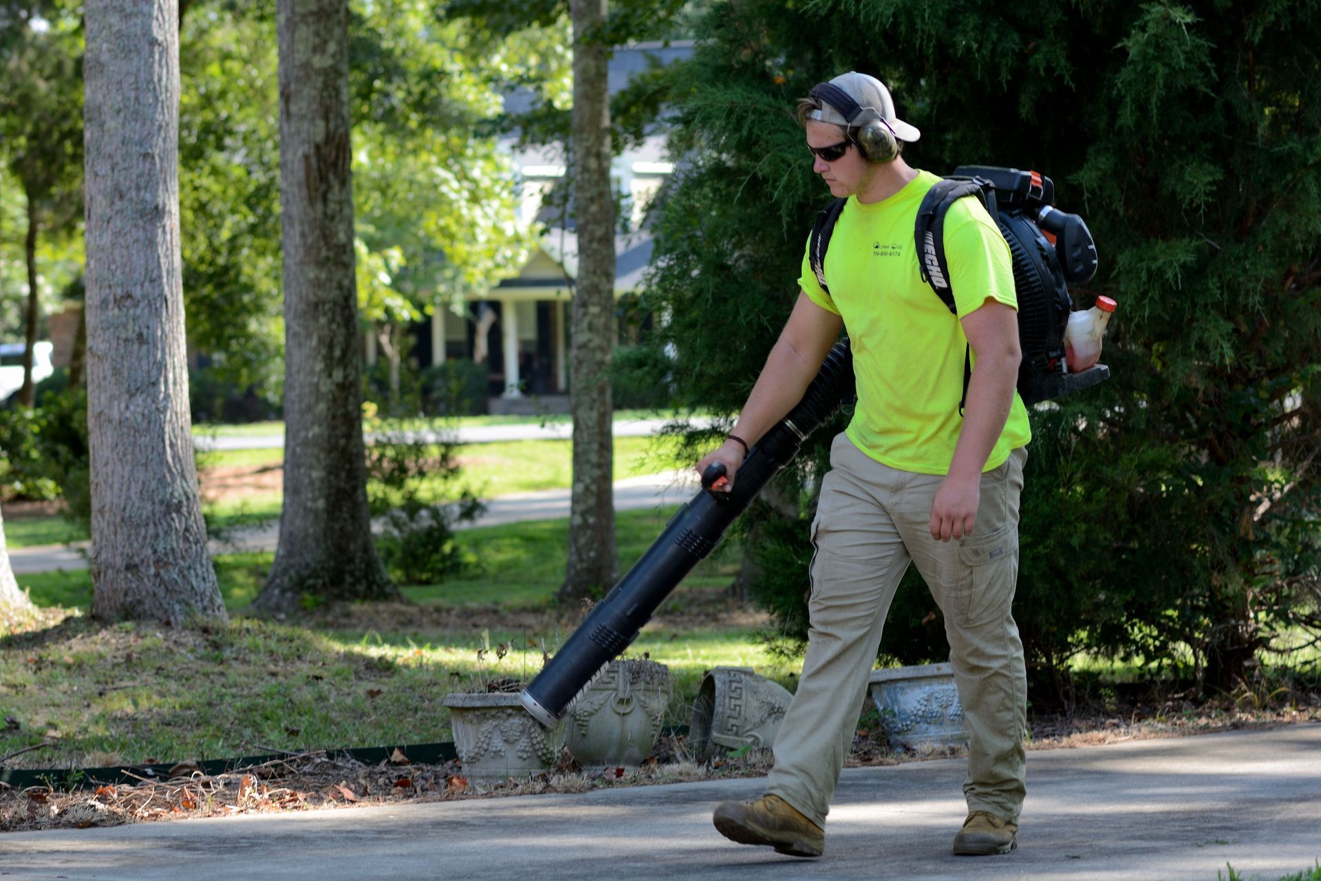 Man using a leaf blower on a sidewalk beside a tree-lined street