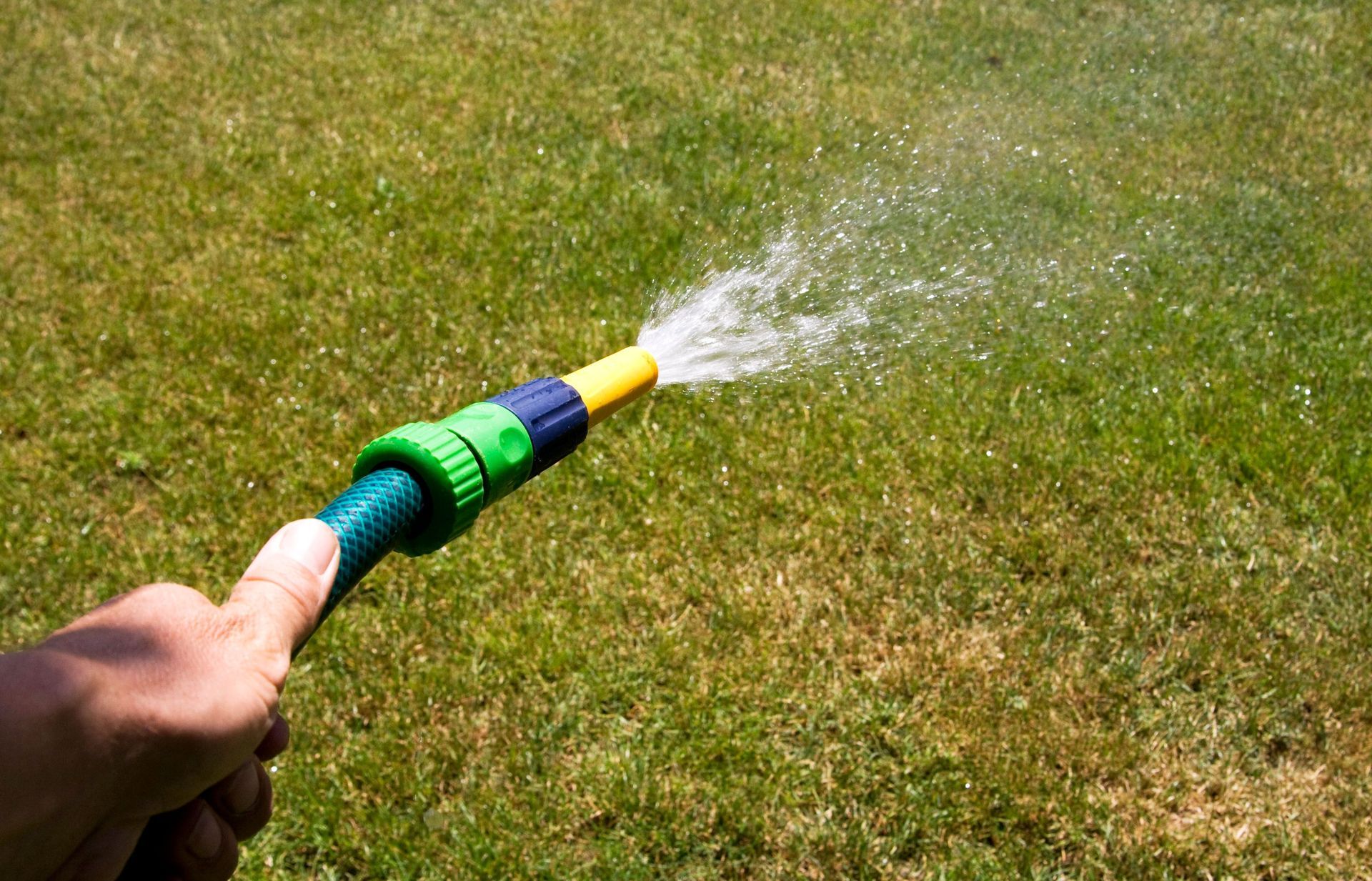 Hand holding a green and yellow garden hose spraying water onto dry grass