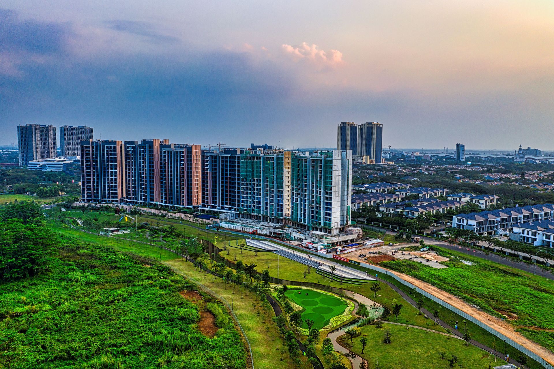 City skyline with high-rise apartments beside green fields and a curving road at dusk