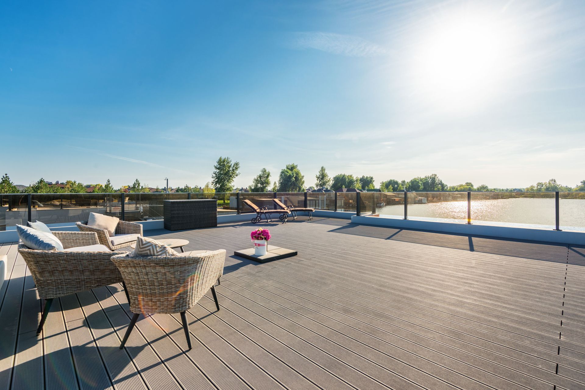 Sunny rooftop deck with lounge chairs, a fire pit, and glass railing overlooking the water.