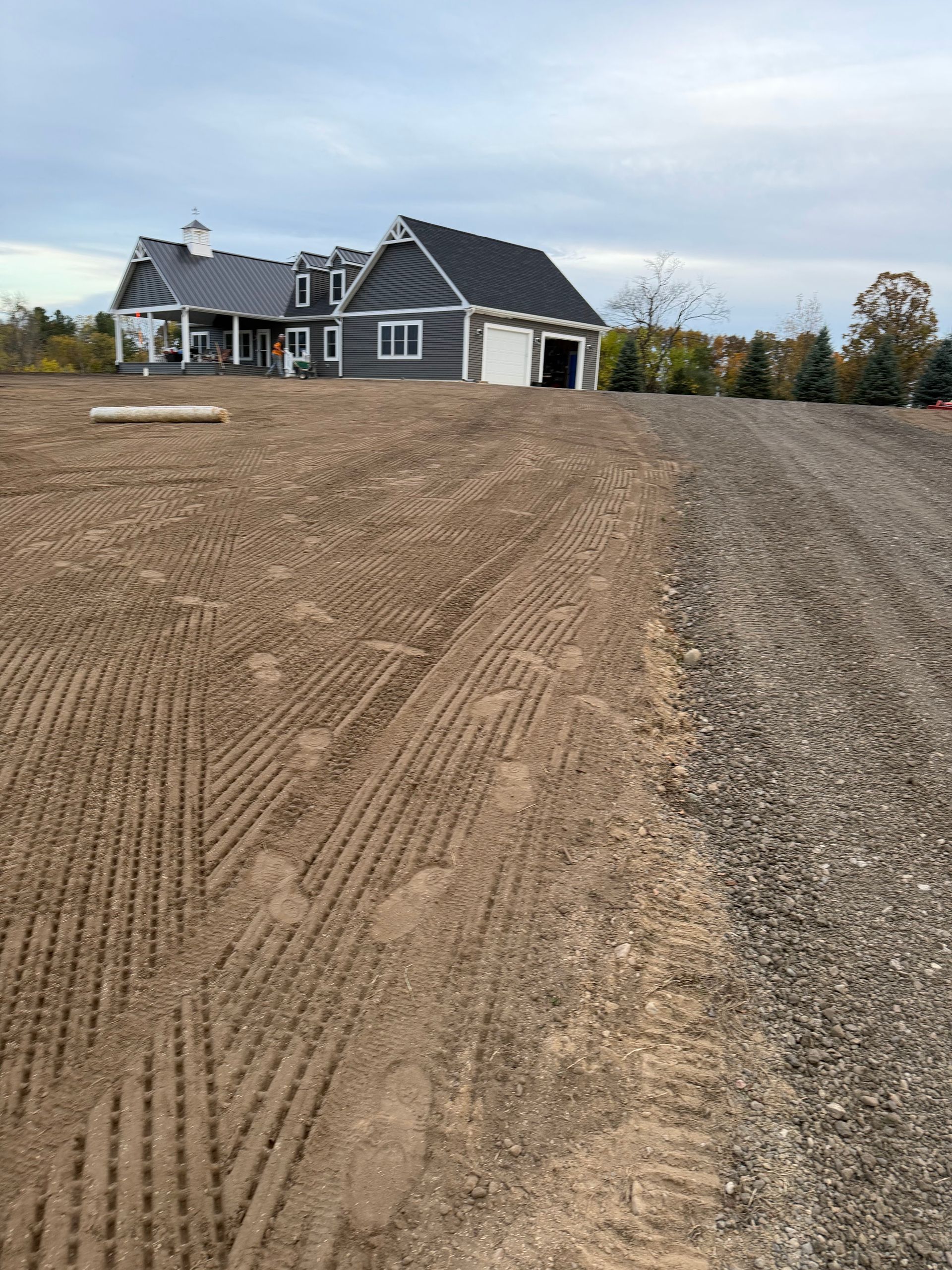 Dirt driveway leading to a house and garage with blue-gray siding under an overcast sky.