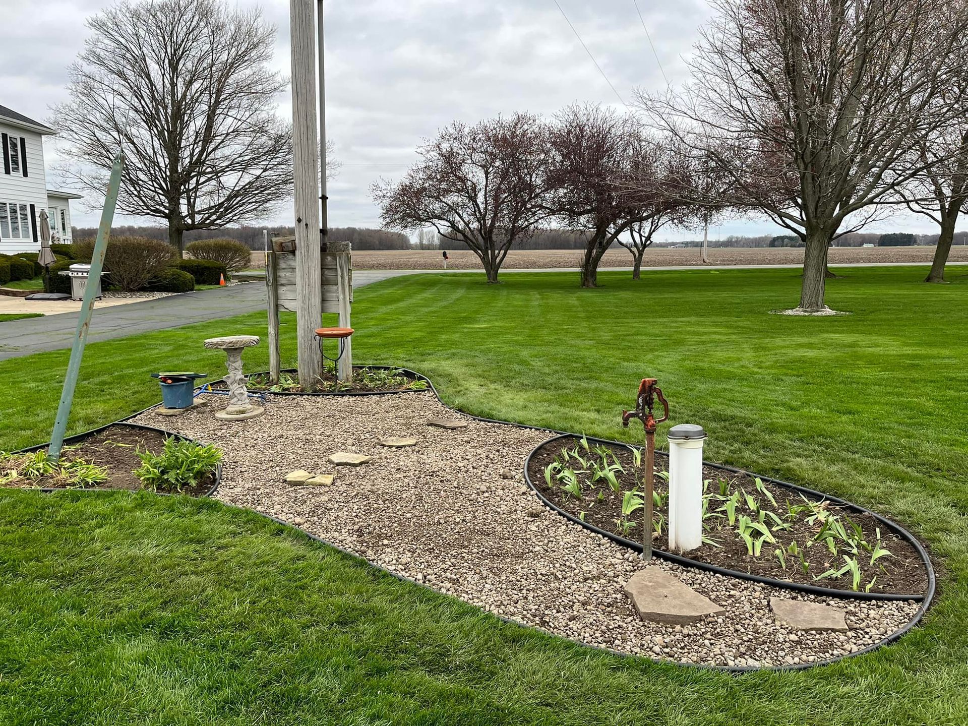 A small garden bed with a flagpole, bird bath, and pump in a grassy yard. Overcast sky.
