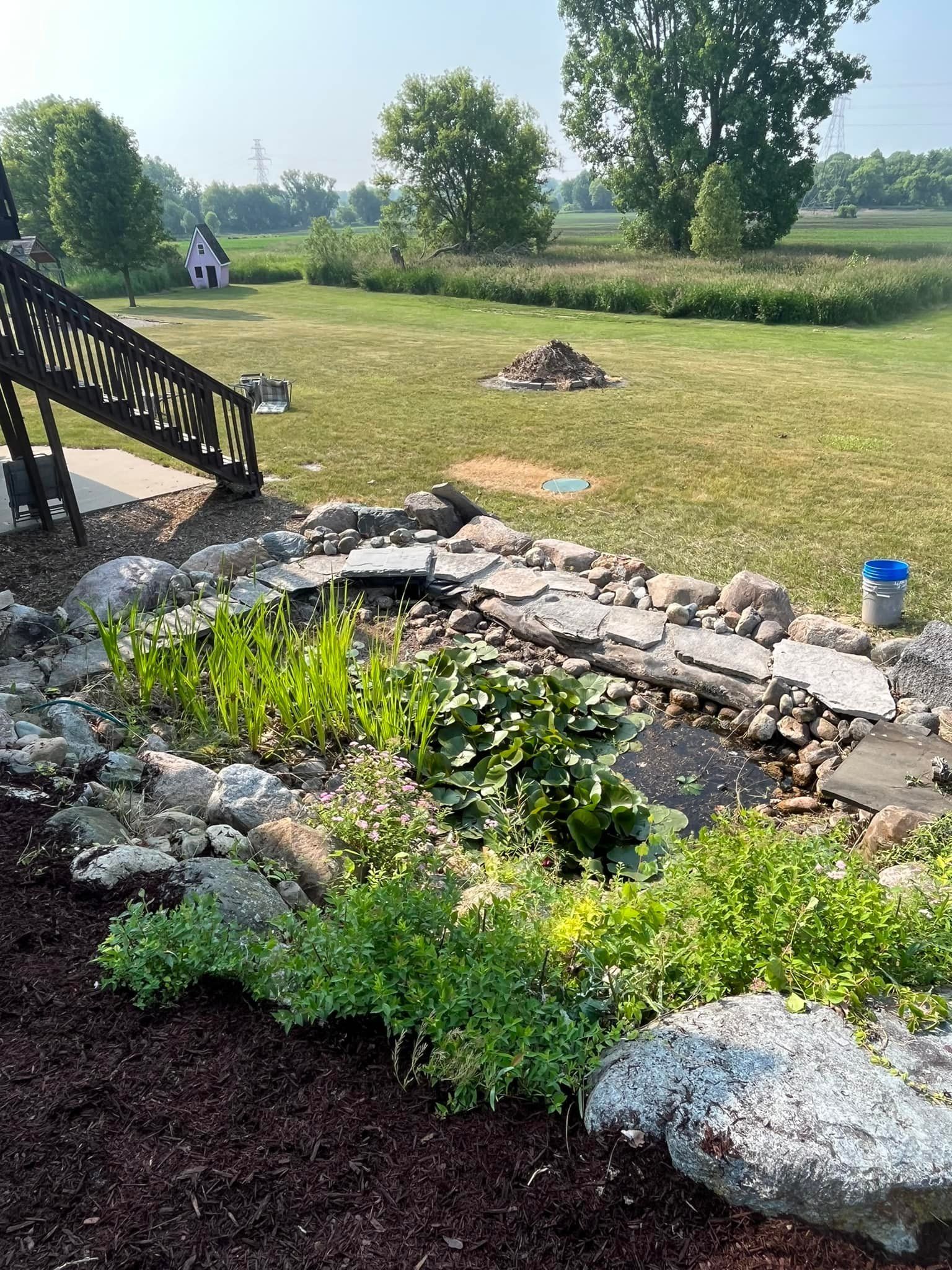 A backyard pond surrounded by rocks and plants, with a wooden deck and grassy field in the background.