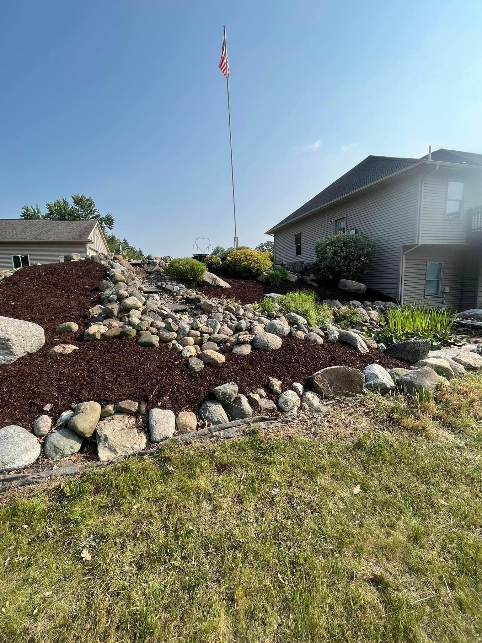 A hillside garden with mulch, rocks, and plants. A house and flag pole are in the background. Sunny day.
