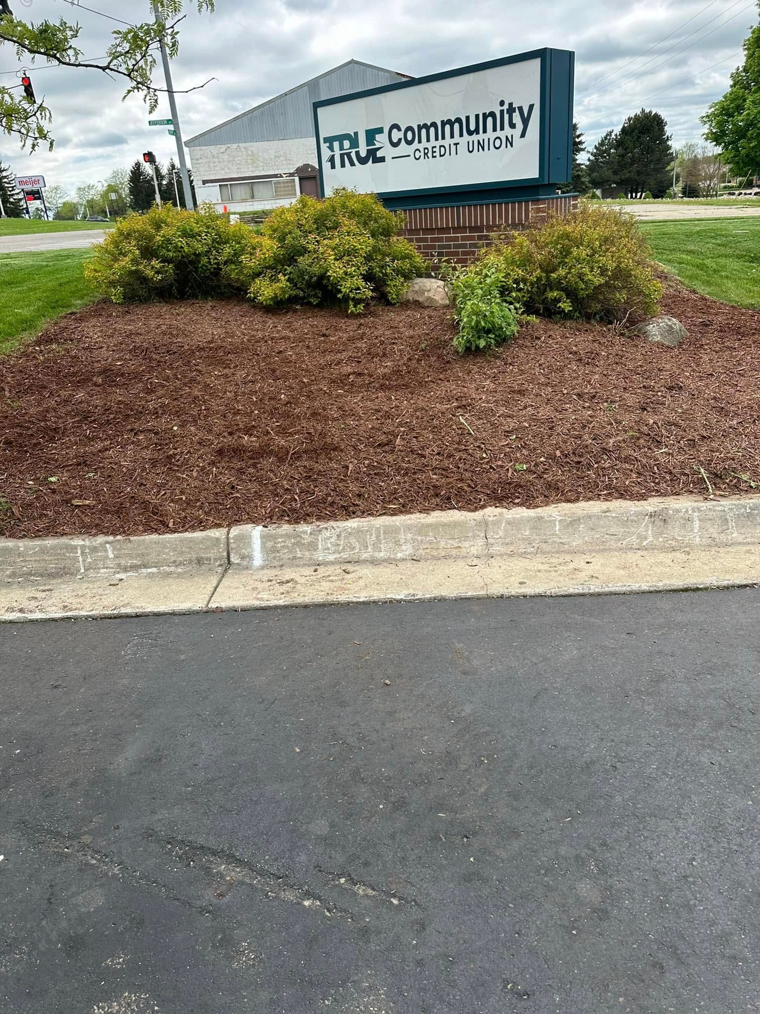 Sign for True Community, brown mulch and shrubs, on a concrete curb, adjacent to a paved road.