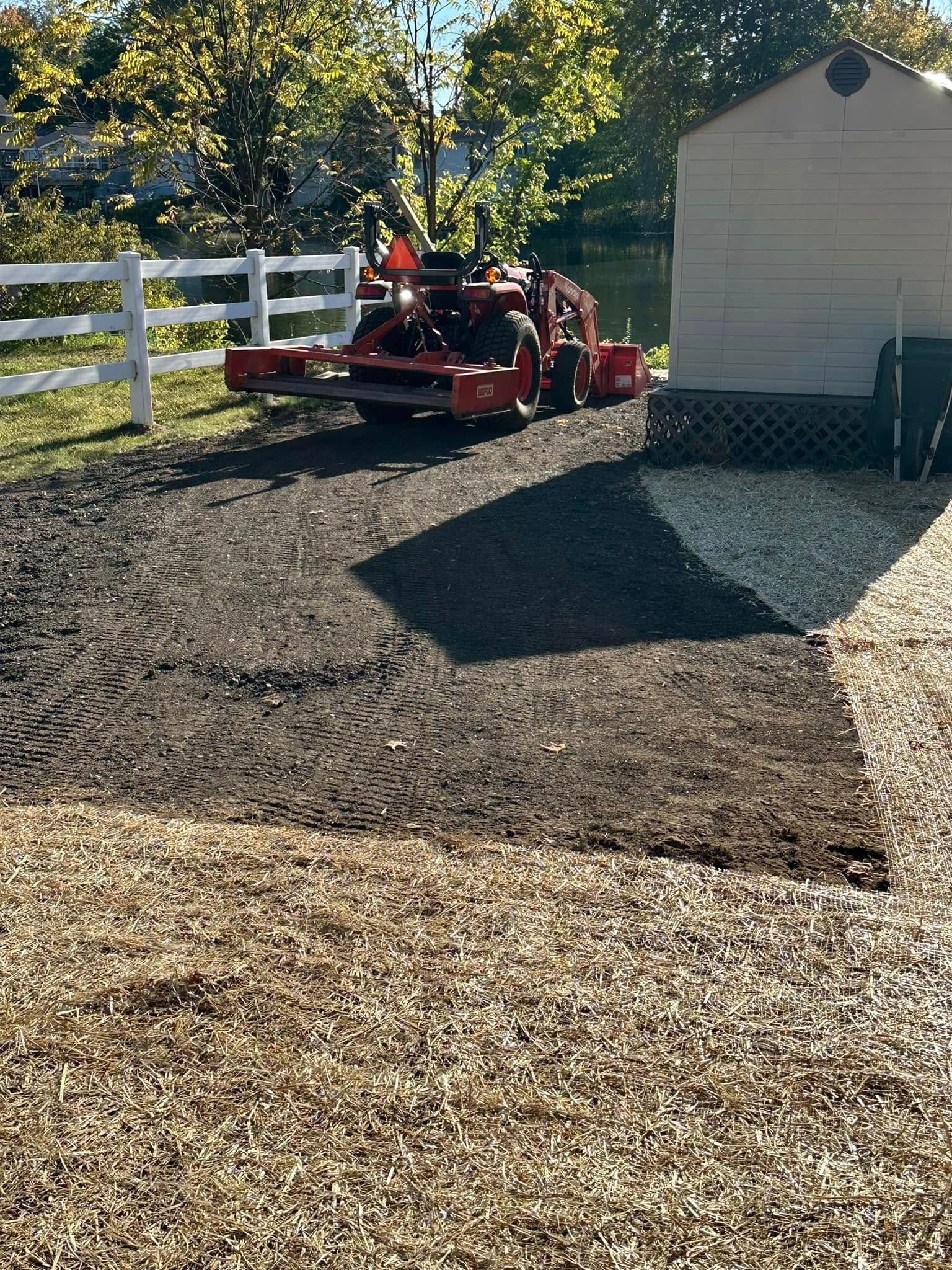 Tractor grading a yard, transitioning from dark soil to lighter gravel. A white fence and shed are visible.