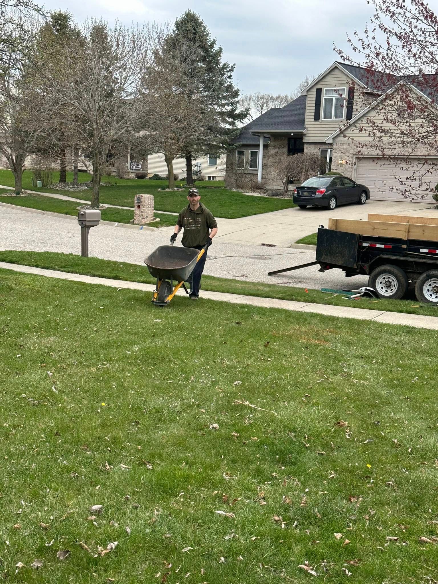 Person pushing a wheelbarrow on a grassy lawn; a trailer is parked nearby.