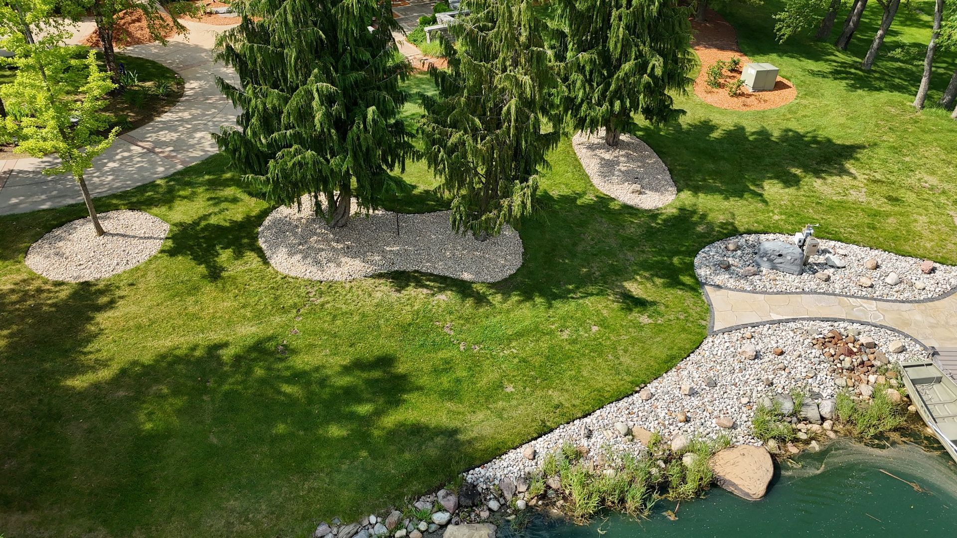 Lush green lawn with stone paths and decorative rocks surrounding trees, near a body of water.