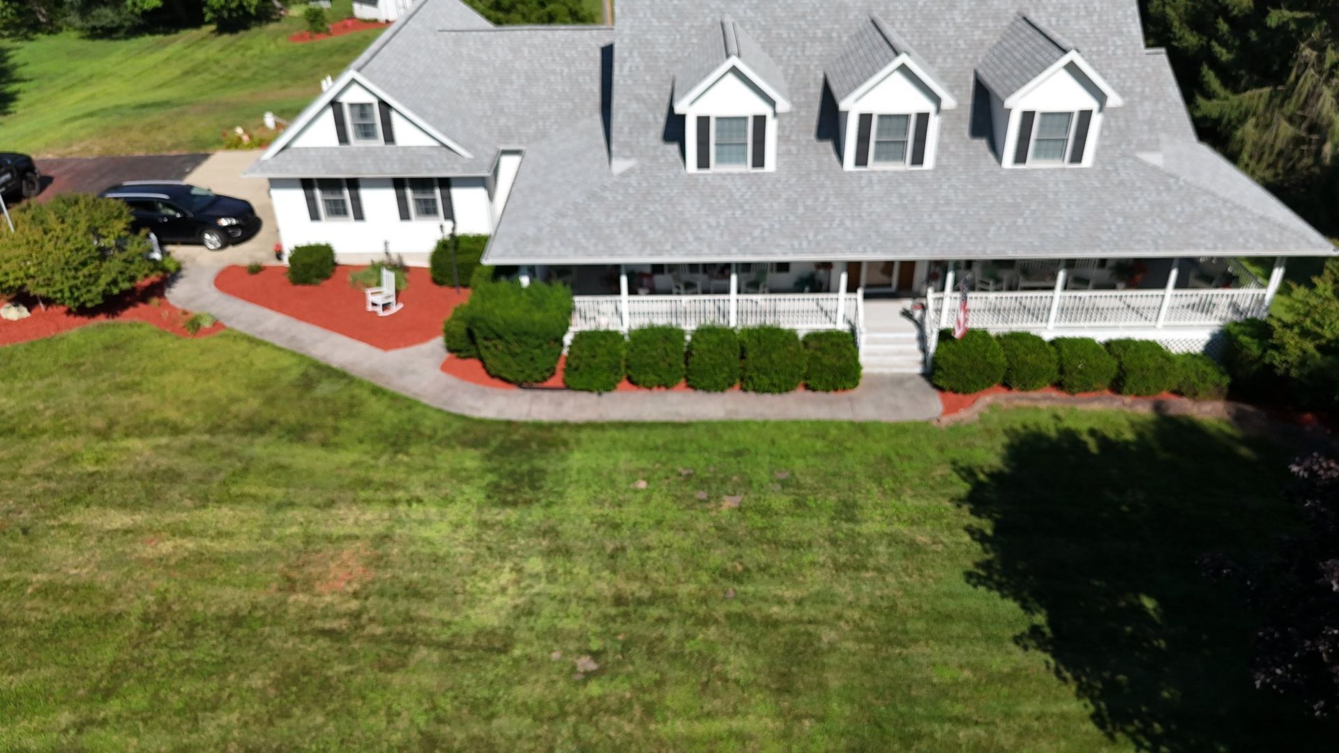 A two-story house with a long porch, three dormers, and a gray roof, in a green lawn.