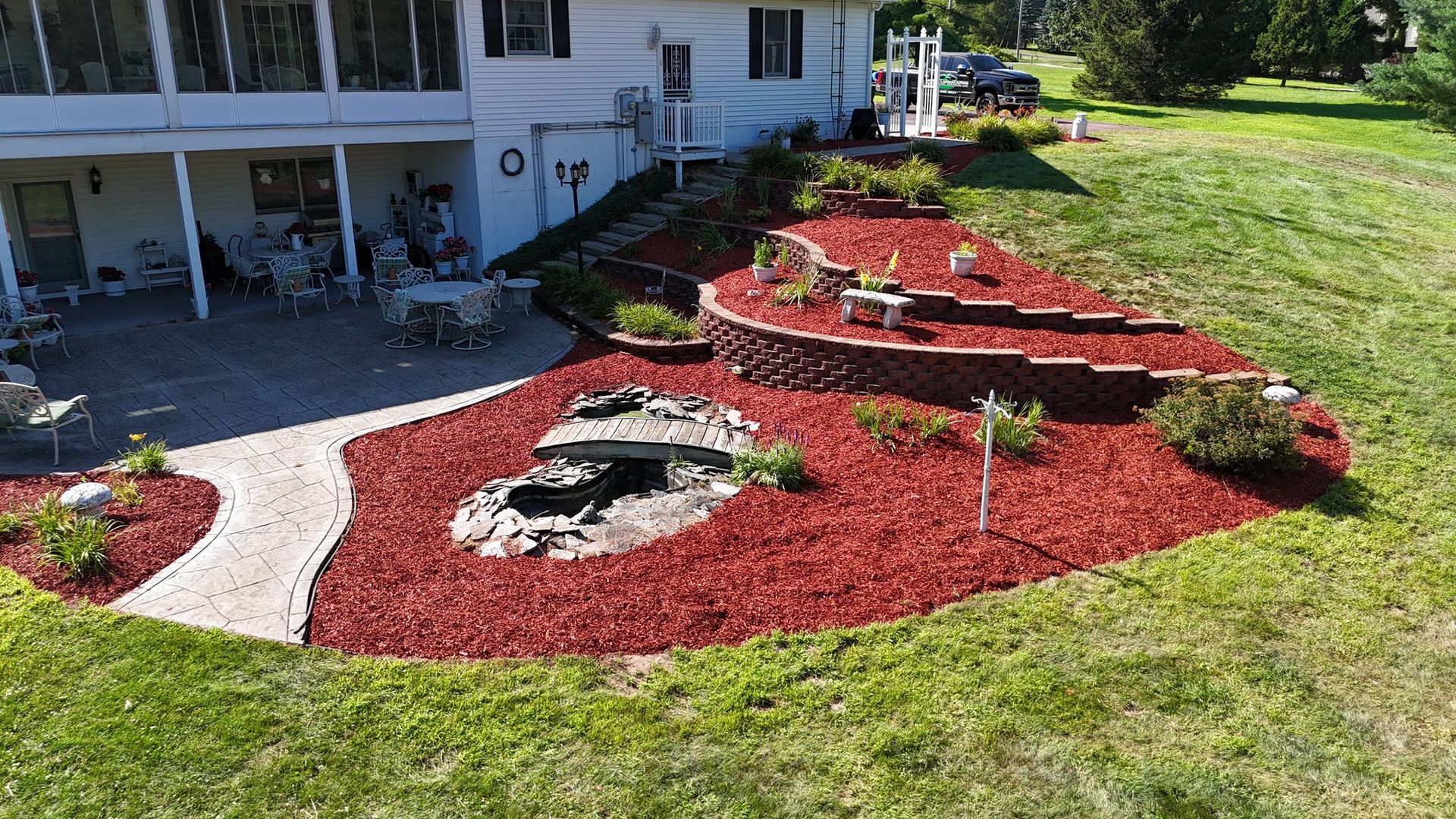 Red mulched landscaping with tiered retaining walls, a curved stone path, and a pond near a two-story white house.