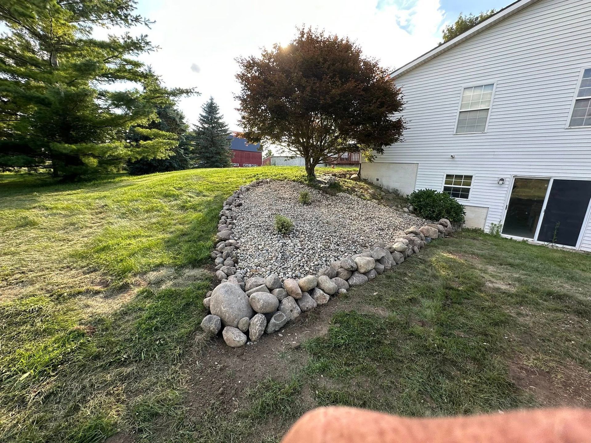 A rock retaining wall borders a sloped lawn with a tree and a house.