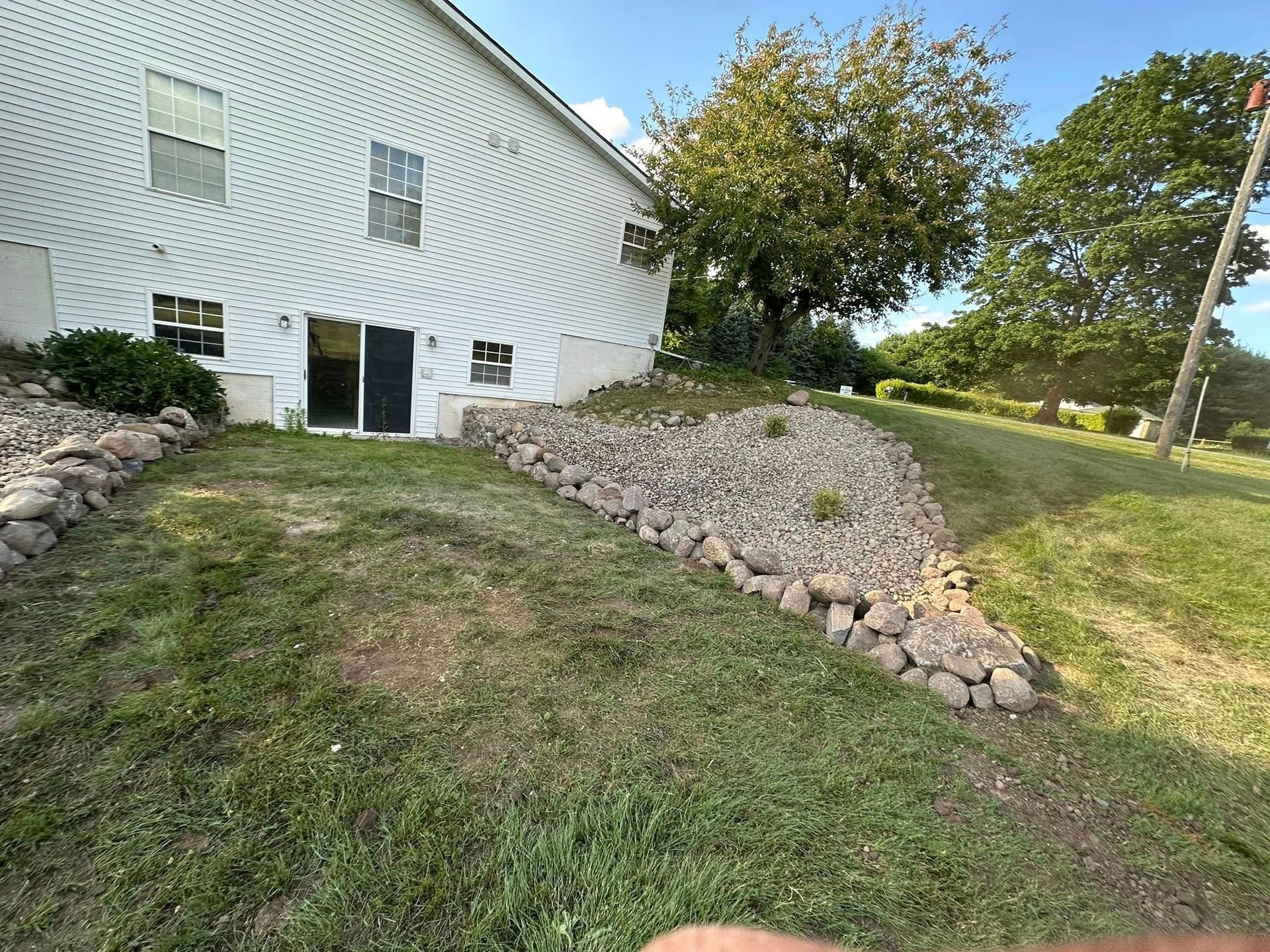 Backyard with rock retaining wall, white house, and green grass on a sunny day.
