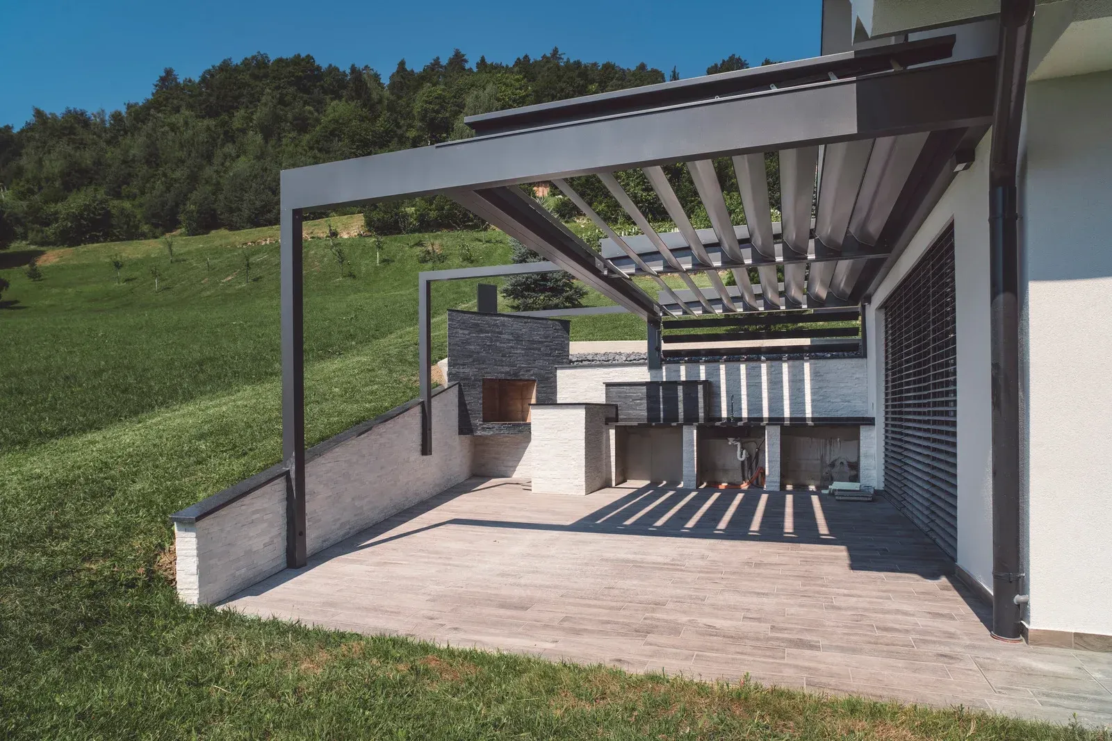 Patio with grey pergola and outdoor kitchen, overlooking a grassy hill.