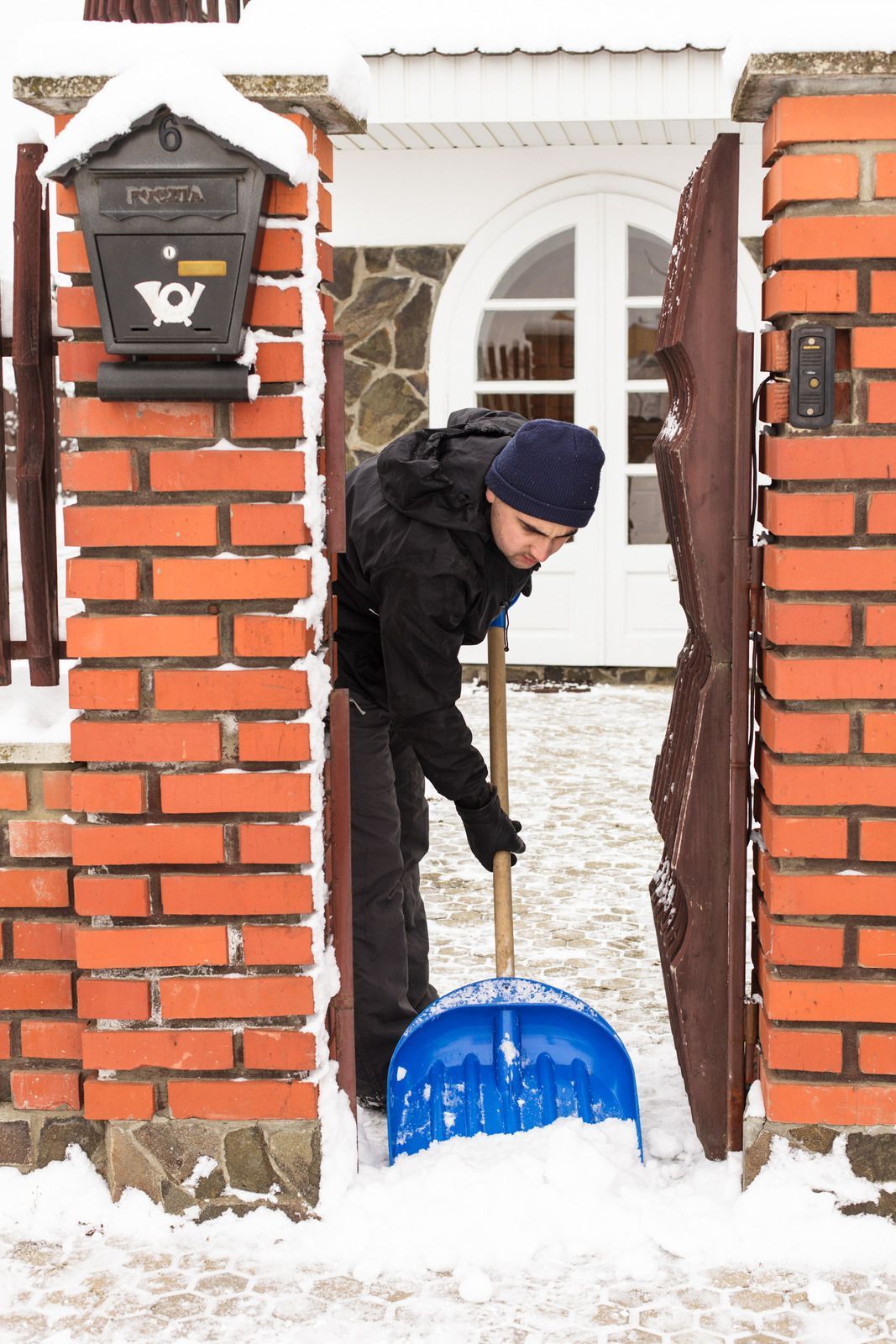 Person shoveling snow from a doorway; brick pillars and a mailbox are on either side.