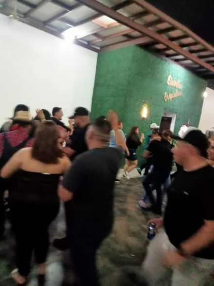 Gente bailando en una habitación muy iluminada con una pared verde; algunos sostienen bebidas.