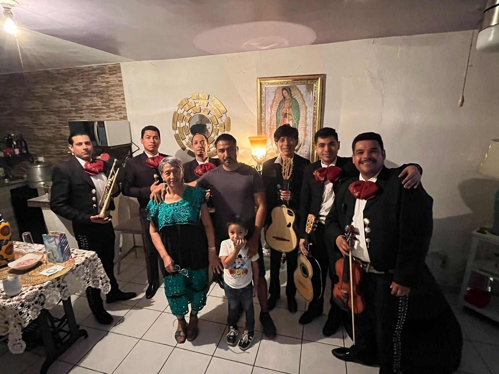 Un mariachi posa con una familia en una casa. Los músicos visten trajes formales y sostienen instrumentos.