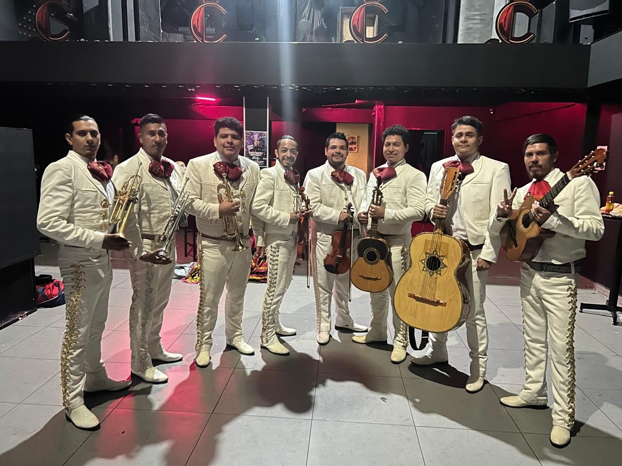 Banda de mariachis con trajes blancos, sosteniendo instrumentos, posando en el interior.