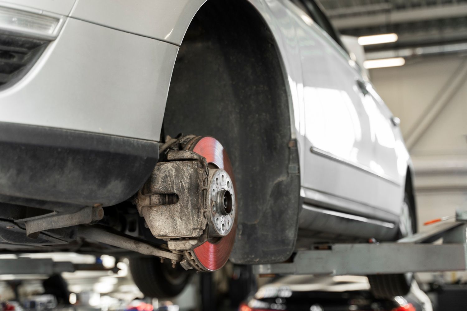 A white car lifted on a garage hoist, highlighting the exposed brake disc and caliper assembly of a removed wheel.