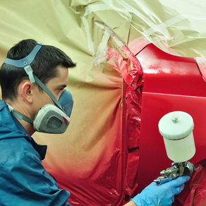 A person in protective gear and a respirator uses a spray gun to paint a car panel red in a workshop.