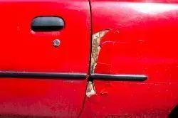 Close-up of a red car door with paint peeling and damaged bodywork near the door seam and trim.