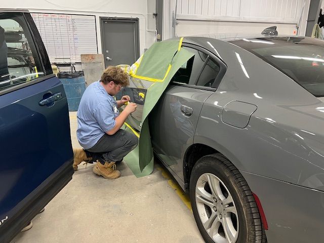 A technician crouches to mask the door of a gray sedan in an automotive repair shop.