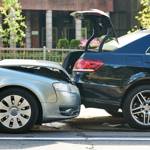 A silver car has crashed into the back of a black car with an open trunk on a paved road.