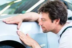 A person in a white uniform inspects the silver body panel of a car, touching it to examine the surface.